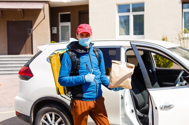 разносчик еды в защитной маске и перчатках с термо рюкзаком возле машины в период карантина
