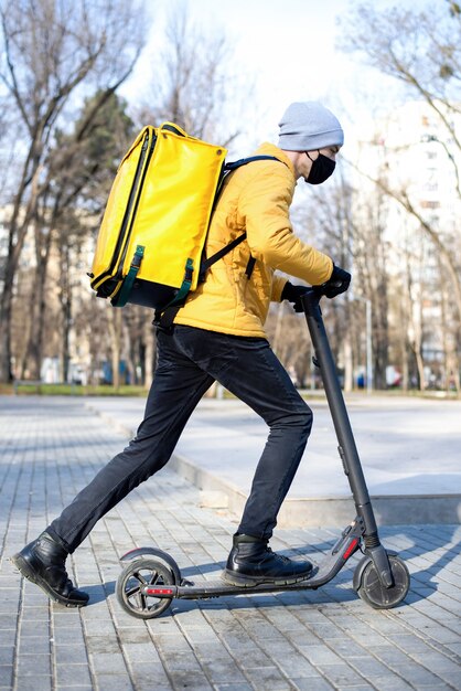 공원에서 스쿠터에 음식 배달 남자. 검은 의료 마스크, 노란색 배낭 및 재킷. 겨울