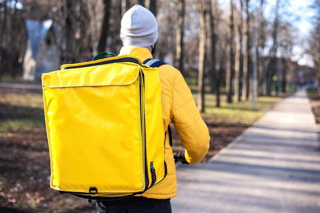 Food delivery man on a scooter in a park. Yellow backpack and jacket. Winter