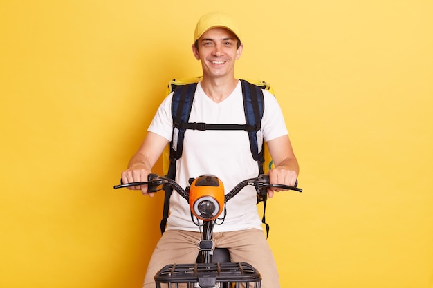 Food delivery Portrait of smiling positive courier guy riding bike with backpack bag delivering meal from takeaway cafe or restaurant isolated over yellow background