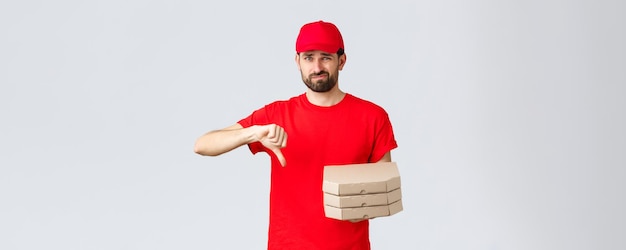 Food delivery quarantine stay home and order online concept Disappointed and reluctant courier in red uniform cap and tshirt dislike pooq quality fastfood thumbdown holding pizza