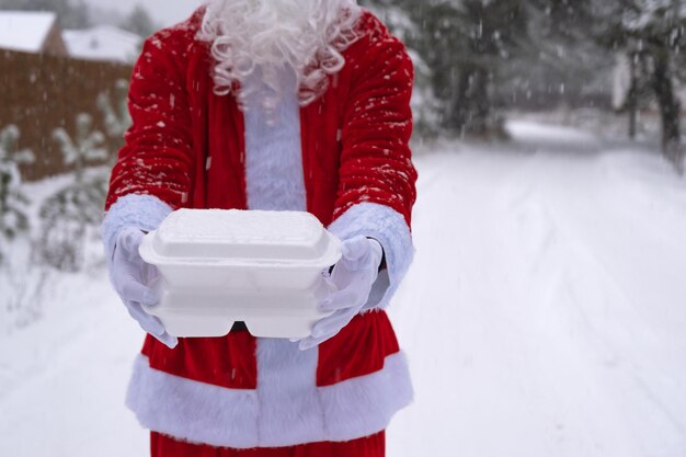 雪の中で屋外のサンタクロースの手に食品配達サービスコンテナー クリスマスイブのプロモーション既製のホットオーダー使い捨てプラスチックボックス新年のケータリングコピースペースのモックアップ