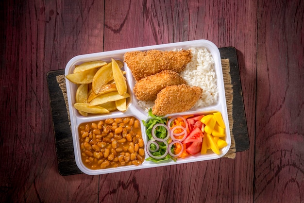 Food delivery, styrofoam lunch box with rice, beans, crispy\
fish fillet, potatoes and salad, top view.