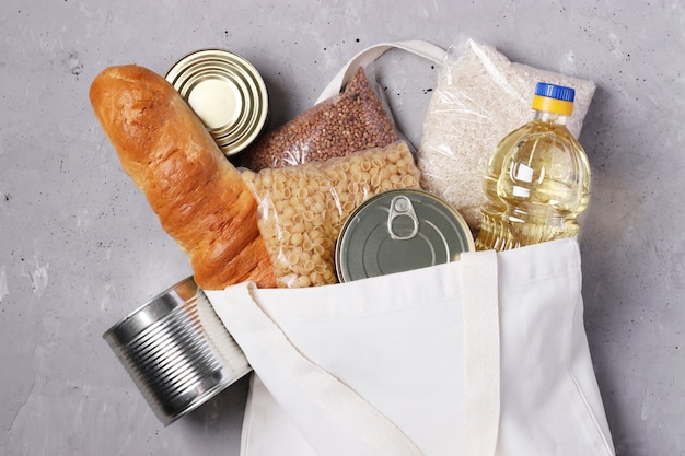 Food delivery. Textile shopping bag with food supplies on gray concrete surface. Rice, buckwheat, pasta, bread, canned food, vegetable oil.