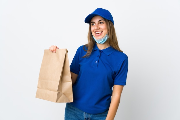 Food delivery woman and protecting from the coronavirus with a mask over isolated white wall looking up while smiling