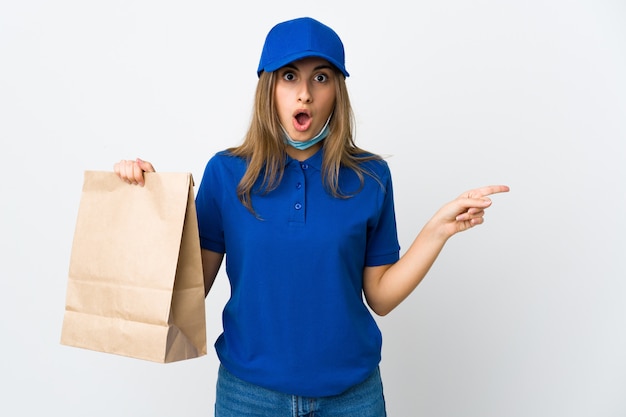 Donna di consegna del cibo e protezione dal coronavirus con una maschera sopra il muro bianco isolato sorpreso e indicando il lato