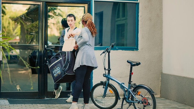 Food delivery worker carrying backpack to deliver fastfood\
order, giving paperbag with lunch meal to client. courier riding\
bike and delivering restaurant food package at office front\
door.