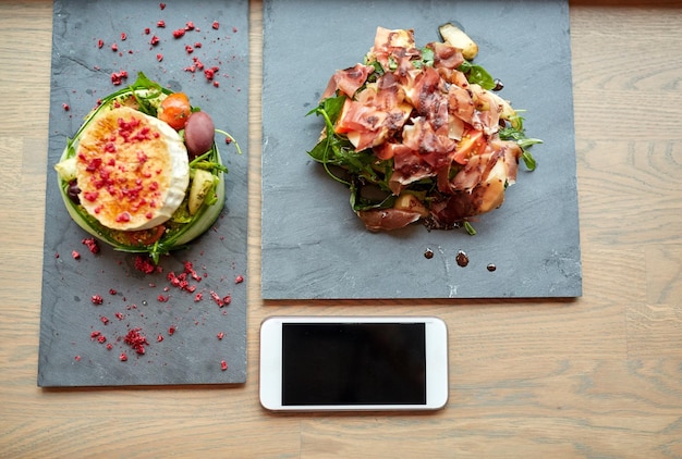 食べ物、食事、テクノロジーのコンセプト – レストランやカフェでスマートフォンを使って石の皿にヤギのチーズと生ハムのサラダを載せる