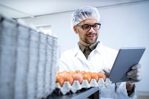 Food factory technologist in white coat hairnet and hygienic gloves controlling eggs production at the food processing plant on tablet computer.