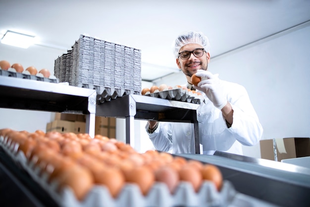 Operaio di fabbrica alimentare in camice bianco e guanti igienici che lavora alla linea di produzione di uova e macchina per l'imballaggio.