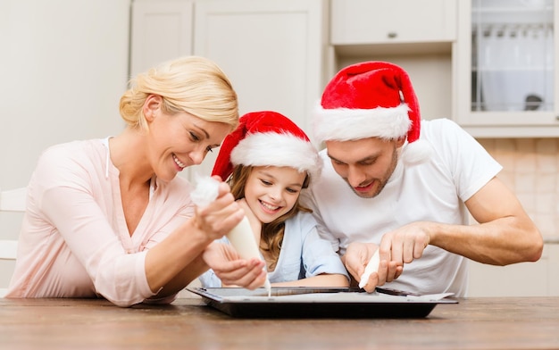 食べ物、家族、クリスマス、幸福と人々の概念-釉薬でクッキーを飾るサンタヘルパー帽子の笑顔の家族