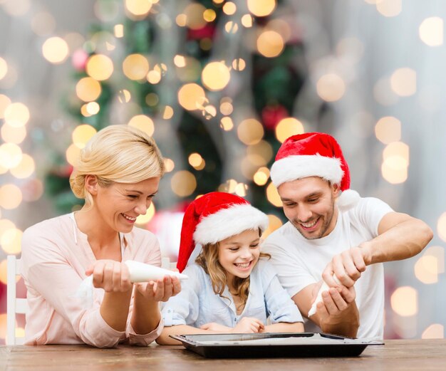 Concetto di cibo, famiglia, felicità e persone - famiglia sorridente in cappelli di babbo natale con glassa e padella che cucinano su sfondo di luci dell'albero di natale