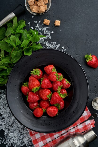 Food fresh ingredients for making lemonade, infused detox water or cocktail. Strawberries, lime, mint, basil, cane sugar, ice cubes and shaker on black stone or concrete background. Top view.