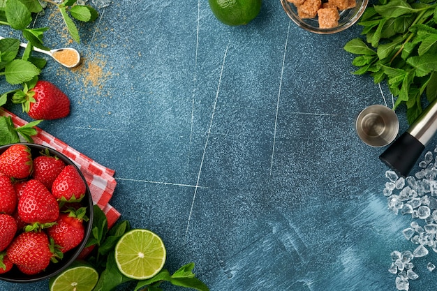 Food fresh ingredients for making lemonade, infused detox water or cocktail. Strawberries, lime, mint, basil, cane sugar, ice cubes and shaker on dark blue stone or concrete background. Top view