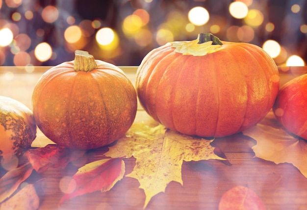 food, halloween, harvest, season and autumn concept - close up of pumpkins and leaves on wooden table over holidays lights