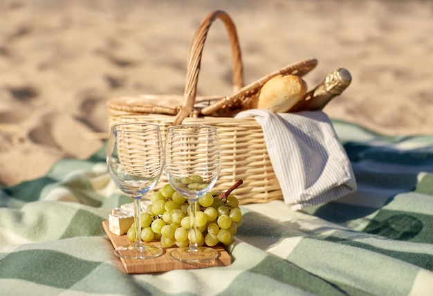 食べ物、休日、お祝いのコンセプト – 夏のビーチにブドウ、ワイングラス、チーズ、シャンパンのボトルを入れたピクニックバスケットの接写