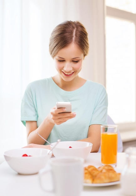 食品、家庭、テクノロジーのコンセプト-スマートフォンでニュースを読んだり、家で朝食をとったりして笑顔の若い女性