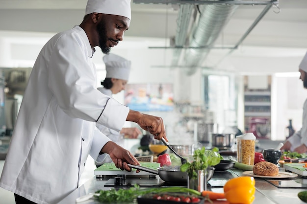 Lavoratore dell'industria alimentare che prepara il pasto nella cucina professionale del ristorante. abile chef che mescola con la spatola in padella mentre cucina un piatto gourmet per il servizio di cena, guarnito con erbe fresche.