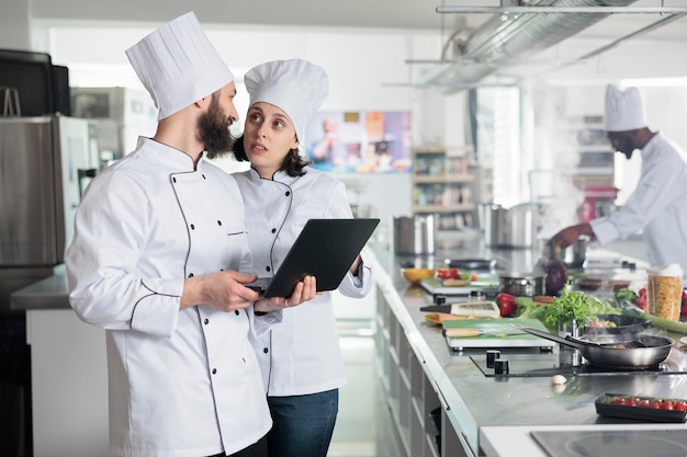 Работники пищевой промышленности, стоящие на профессиональной кухне ресторана, используют компьютер, чтобы выбрать блюдо для ужина. Шеф-повара ищут рецепты изысканной кухни на компьютере. Концепция приготовления