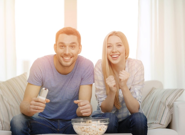 food, love, family, sports, entretainment and happiness concept - smiling couple with popcorn cheering sports team at home