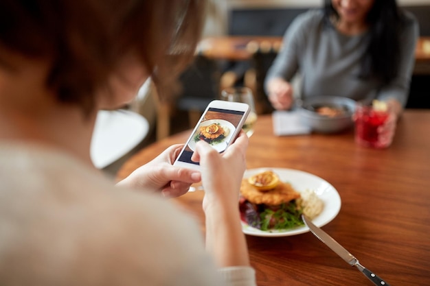 Foto cibo, nuova cucina nordica, tecnologia e concetto di persone - donne con smartphone che mangiano filetto di pesce al pane con salsa tartara e insalata di pomodoro di barbabietole al forno per cena al ristorante