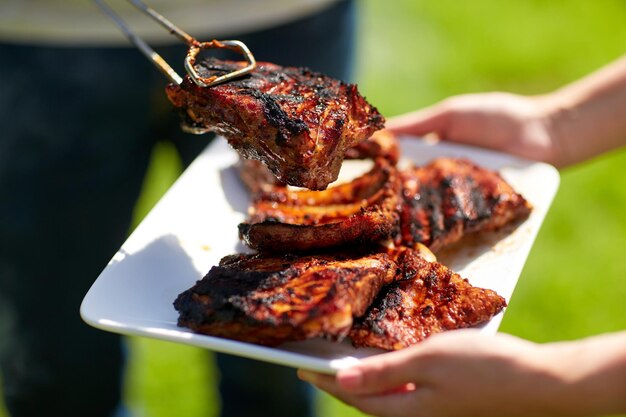 food, people, eating and cooking concept - man with tongs putting barbecue meat on plate at summer party