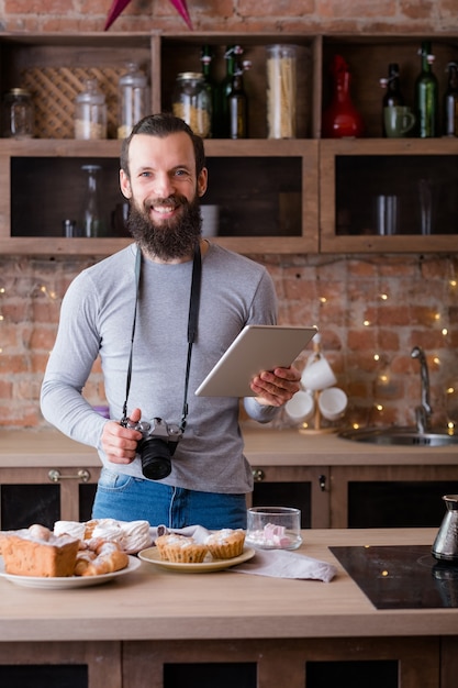 Food photographer. Blogging business. Smiling man with tablet and camera. Cakes and pastries assortment around.