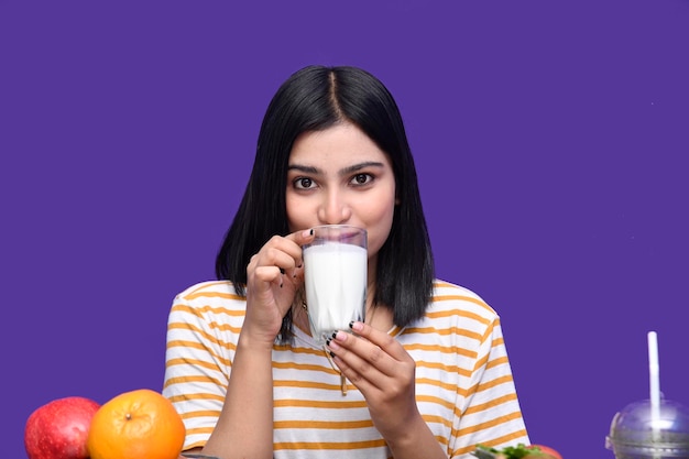 Foodie girl sitting at fruit table drinking milk over purple background indian pakistani model