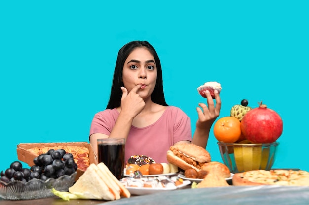 Photo foodie girl sitting at fruit table eating cupcake over blue background indian pakistani model