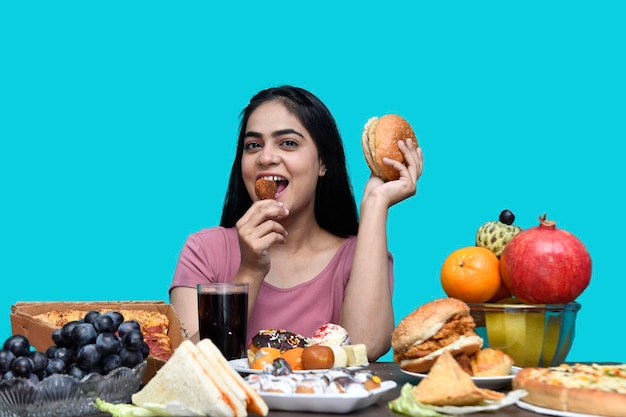 Photo foodie girl sitting at fruit table holding burger and eating fried chicken indian pakistani model