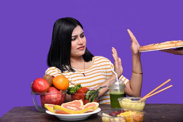 Photo foodie girl sitting at fruit table with open hands stop sign to pizza indian pakistani model