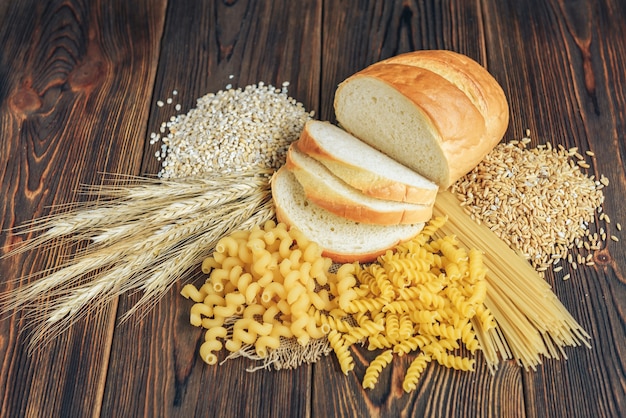 Foods high in carbohydrate on wooden background. Loaf, pasta, pearl barley and oats.