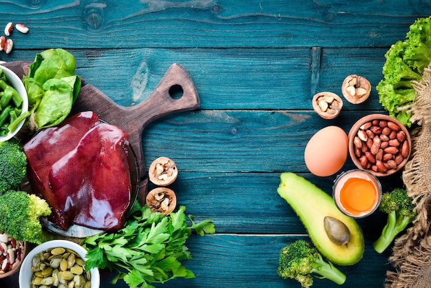 Продукты, содержащие натуральный витамин B9 Печень, авокадо, брокколи, шпинат, петрушка, фасоль, орехи на синем фоне Вид сверху
