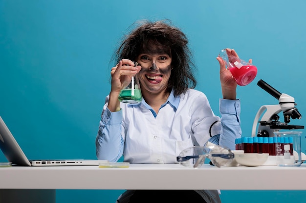 Foolish scientist with funny face expression handling beakers filled with serum. Mad silly looking crazy lab worker sitting at desk while having glass jars filled with experimental liquid compounds.