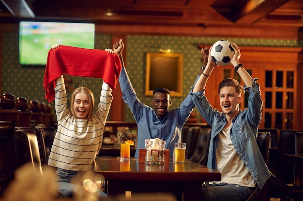 Football fans with red scarf watching game translation, friends in bar. Group of people relax in pub, night lifestyle, friendship, sport celebration