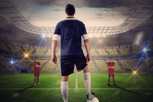 Football player in blue with ball facing opposition against large football stadium with lights