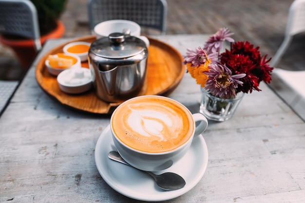 In the foreground a cup of coffee with foam a spoon and a plate\
tray with accessories for tea