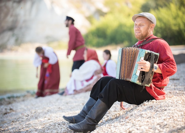 In the foreground, a man in a Russian folk costume sits and plays the accordion against a beautiful landscape and his friends, close up