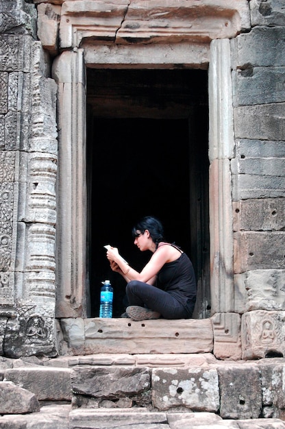 2009年4月12日にシェムリアップカンボジアのシェムリアップにあるアンコールワットの古代遺跡の建物で写真を撮るために、外国人の若い女性旅行者が訪問し、肖像画のポーズをとってリラックスして休憩をとる