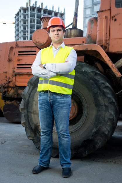 Foreman in hardhat posing next to bulldozer with crossed hands