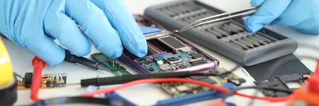 Foreman in rubber gloves repairing mobile phone with tweezers in workshop closeup