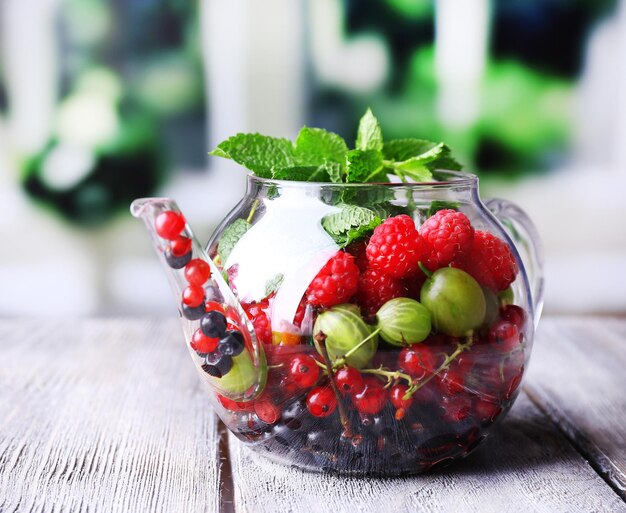 Forest berries and mint leaves in glass teapot on wooden table on bright background