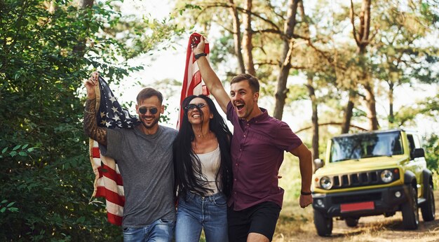 In the forest. Friends have nice weekend outdoors near theirs green car with USA flag.