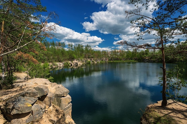 A forest lake with rocky shores and coniferous trees with a blue sky and white clouds reflected in the water