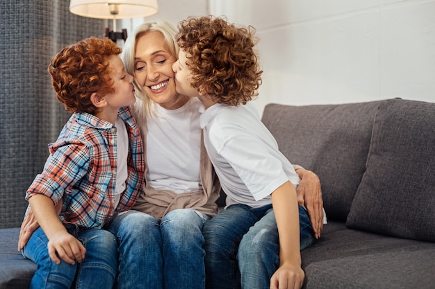 Photo forever thankful. curly haired boys kissing their granny cheeks while all sitting on a sofa and expressing their love to her at home.