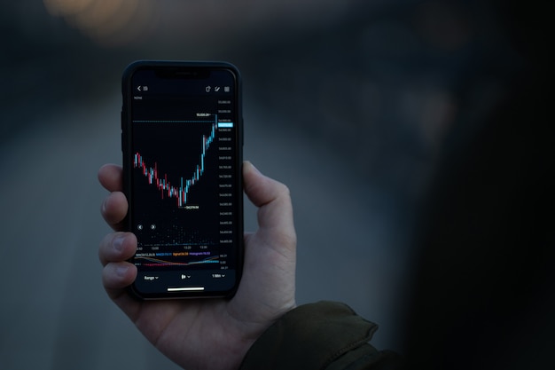 외환 거래. 남성의 손에 화면에 촛대 차트가있는 스마트 폰을 들고, 상인은 금융 뉴스를 읽고 야외에서 stnading하는 동안 모바일 앱에서 실시간 외환 시장 데이터를 확인합니다.