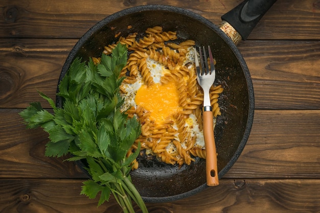 Fork in a frying pan with pasta and egg with parsley on a wooden table