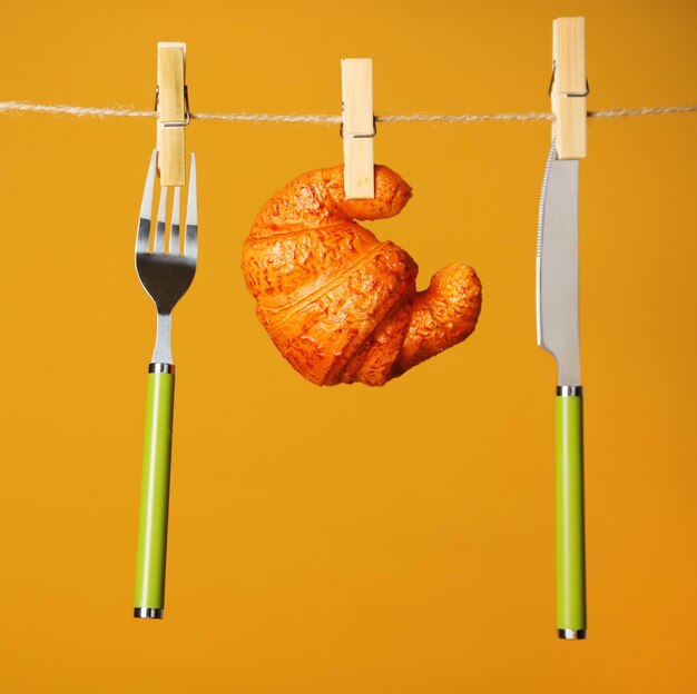 Fork knife and croissant hanging from clothesline on color background