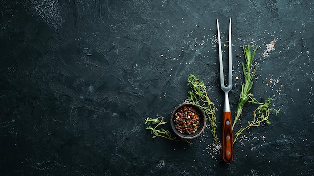 Forchetta per carne, spezie ed erbe aromatiche su un tavolo di pietra nera vista dall'alto spazio libero per il testo