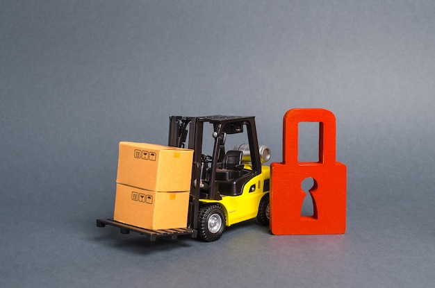 Photo forklift truck carries a cardboard box next to red padlock embargo trade wars no delivery restriction on the importation of goods proprietary for business inability to sell products ban import